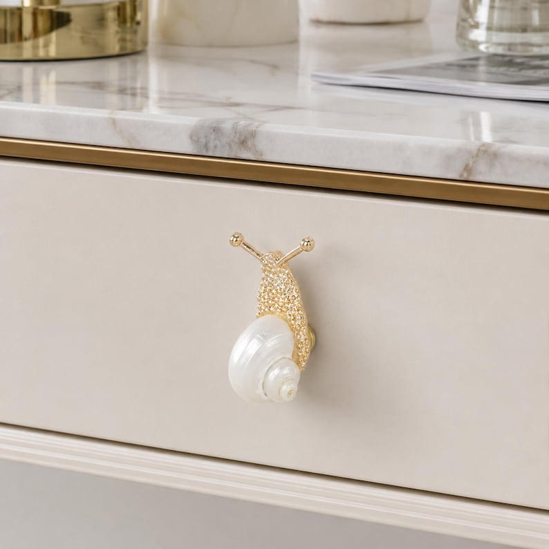 May include: A snail-shaped drawer knob featuring a textured gold-tone metal body with two antennae and a smooth, iridescent white shell. The decorative hardware is mounted on a light beige drawer front with a horizontal gold trim detail.