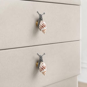 May include: Two drawer knobs shaped like snails with textured dark gray metal bodies and spiraled, tan and white patterned seashell tops. Each snail knob is mounted on a small, circular gold-toned base attached to a light beige drawer front.