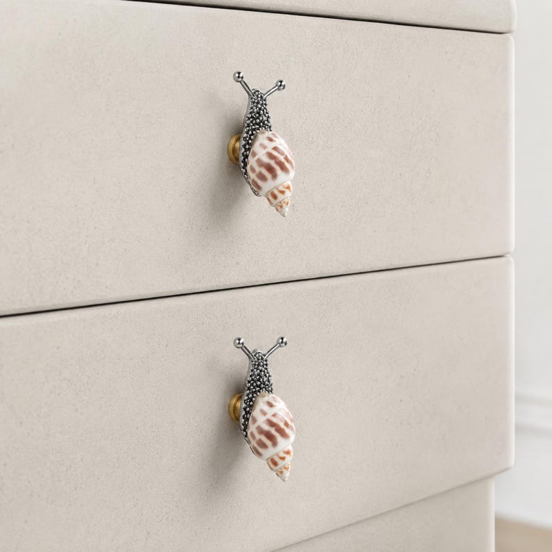 May include: Two drawer knobs shaped like snails with textured dark gray metal bodies and spiraled, tan and white patterned seashell tops. Each snail knob is mounted on a small, circular gold-toned base attached to a light beige drawer front.