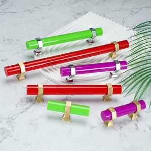 May include: A collection of six cylindrical cabinet pulls in various lengths and colors, including bright green, red, and purple. Each pull features metallic mounting brackets in either silver or gold tones, designed for attachment to furniture.