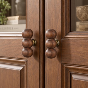 Puede incluir: Dos pomos de armario a juego hechos de madera de color marrón oscuro, cada uno con una pila vertical de tres formas esféricas. Los pomos están unidos a una puerta de armario de madera con una base de metal color latón, mostrando una veta de madera natural.