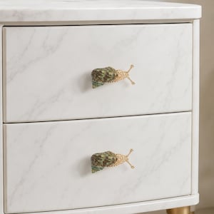 May include: A white marble-patterned two-drawer cabinet featuring two decorative drawer pulls shaped like snails. Each snail pull has a textured green and brown shell with a gold-toned metallic body and two protruding antennae.