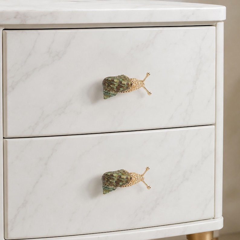 May include: A white marble-patterned two-drawer cabinet featuring two decorative drawer pulls shaped like snails. Each snail pull has a textured green and brown shell with a gold-toned metallic body and two protruding antennae.