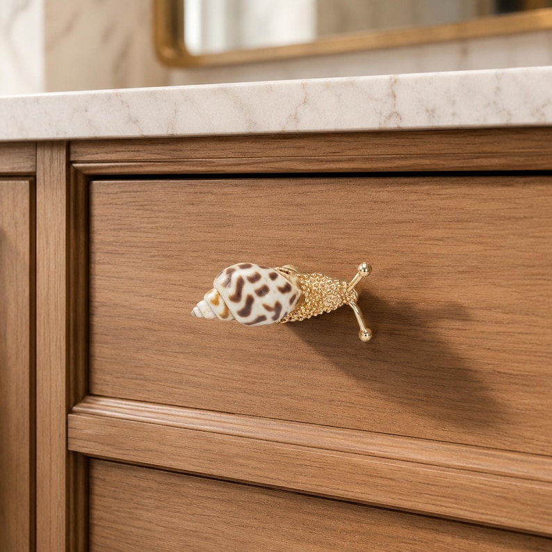 May include: A cabinet knob shaped like a snail, featuring a natural brown and white patterned seashell for the shell and a textured gold-toned metal body with two antennae extending outward, mounted on a wooden drawer front.
