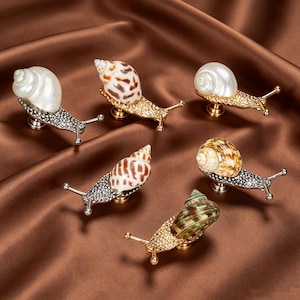 May include: A collection of six snail-shaped cabinet knobs featuring real seashells as shells. The bodies are crafted from textured metal in gold and silver tones, each with two protruding antennae and a central mounting base for furniture.
