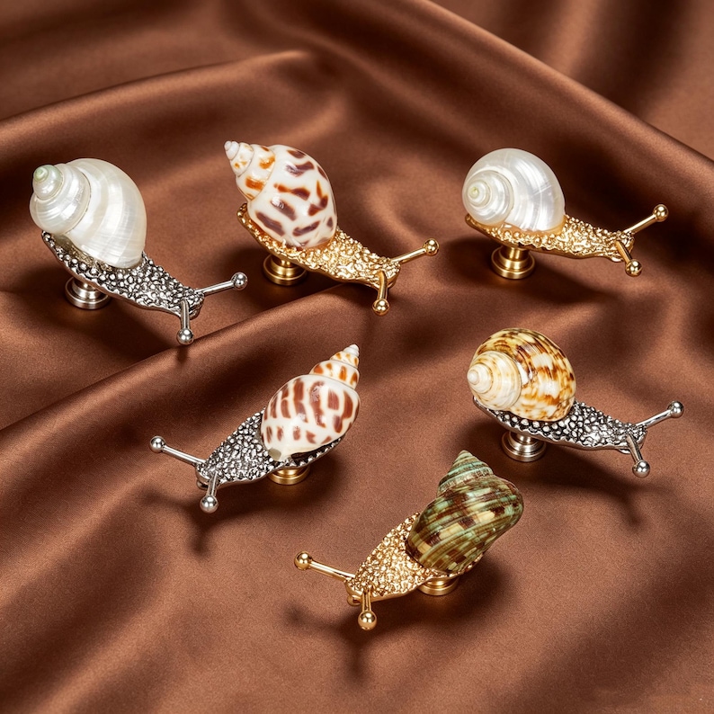 May include: A collection of six snail-shaped cabinet knobs featuring real seashells as shells. The bodies are crafted from textured metal in gold and silver tones, each with two protruding antennae and a central mounting base for furniture.