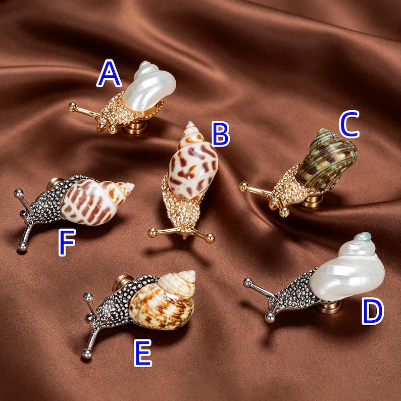May include: Six snail-shaped cabinet knobs labeled A through F. Each knob features a metallic body encrusted with small crystals, paired with a unique natural seashell. The snails have two protruding antennae and a mounting post on the underside.