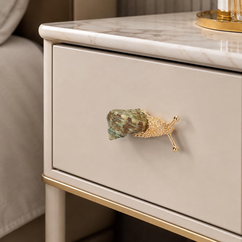 May include: A drawer knob shaped like a snail with a textured gold metal body and a natural green and brown spiral shell. The knob is attached to a smooth, light beige drawer front on a piece of furniture with a white marble top surface.
