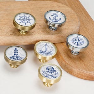 Puede incluir: Un juego de seis pomos redondos para armarios con diseños náuticos en azul y blanco. Los pomos tienen acabados metálicos en tonos latón y plata. Los diseños incluyen un timón de barco, un ancla, un faro, un velero, una brújula y una calavera.
