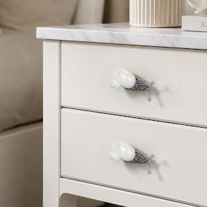 May include: Two drawer pulls shaped like snails attached to a white nightstand. Each pull features a glossy, iridescent white seashell as the snail shell and a textured, dark metallic body with two thin, silver-toned antennae extending outward.