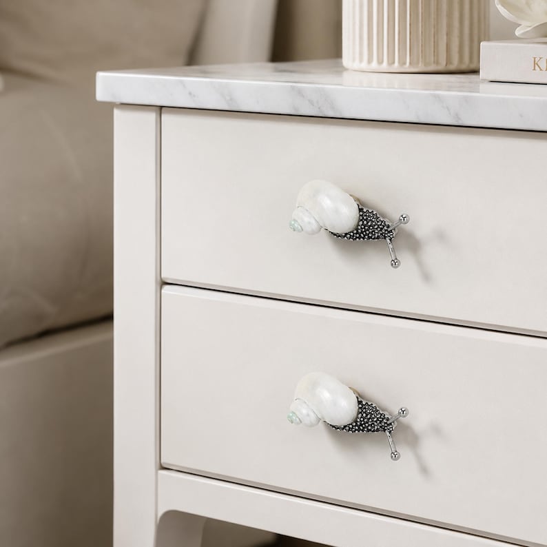 May include: Two drawer pulls shaped like snails attached to a white nightstand. Each pull features a glossy, iridescent white seashell as the snail shell and a textured, dark metallic body with two thin, silver-toned antennae extending outward.