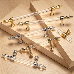 Könnte beinhalten: Eine Sammlung von Schrankgriffen und einem einzelnen Knopf mit zylindrischen Stangen aus klarem Acryl sowie metallischen Endkappen und Montagepfosten. Die Beschläge sind in polierten Gold- und Silberausführungen erhältlich.