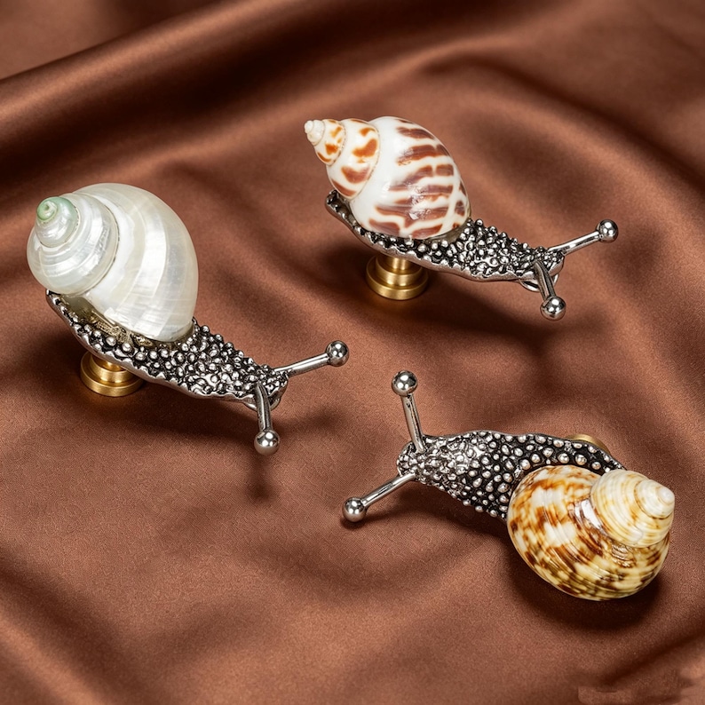 May include: Three snail-shaped cabinet knobs featuring textured, dark metallic bodies with extended antennae and brass-colored mounting bases. Each knob is topped with a unique, polished natural seashell, showcasing varied brown and white patterns.