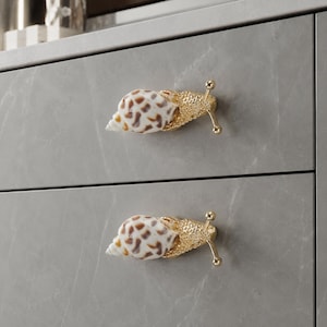 May include: Two drawer knobs shaped like snails, featuring a white and brown spotted seashell for the shell and a textured gold-tone metal body with two antennae. The knobs are mounted on a gray cabinet with a subtle white marble-like pattern.