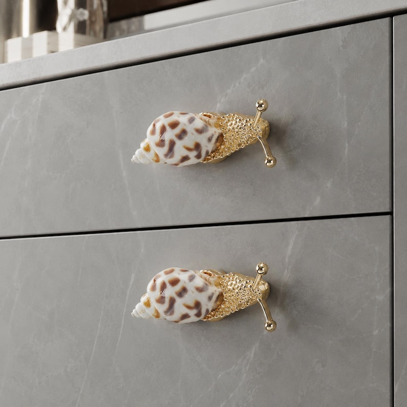 May include: Two drawer knobs shaped like snails, featuring a white and brown spotted seashell for the shell and a textured gold-tone metal body with two antennae. The knobs are mounted on a gray cabinet with a subtle white marble-like pattern.