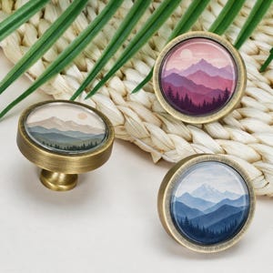 Puede incluir: Tres pomos redondos para armarios con acabado en latón cepillado. Cada pomo presenta una inserción de cristal circular que muestra un paisaje de montaña diferente: uno con tonos rosas y morados, otro con tonos tierra y otro con tonos azules.