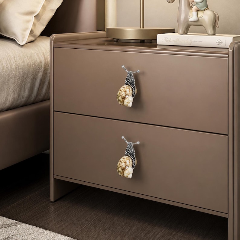 May include: Two drawer pulls shaped like snails with dark, speckled bodies and textured, tan and cream-colored spiral shells. The snail-shaped knobs are mounted on the front of a brown two-drawer nightstand, serving as functional handles.