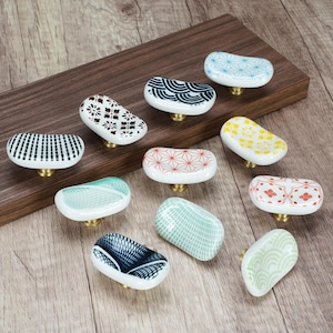 May include: A collection of ten oval-shaped ceramic cabinet knobs featuring various patterns including geometric lines, waves, and floral motifs in colours such as blue, red, yellow, green, and black, each mounted on a small brass-coloured metal base.