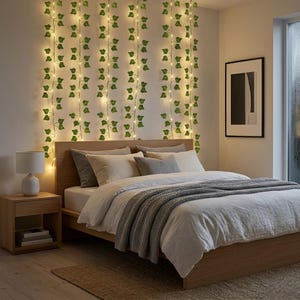 Peut inclure: Une guirlande de feuilles de lierre artificielles vertes entrelacées de petites lumières LED blanc chaud, suspendue verticalement contre un mur derrière un cadre de lit en bois. La guirlande lumineuse présente un feuillage vert réaliste.
