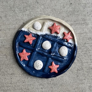May include: A ceramic, round, nautical-themed decorative piece. It features a blue grid pattern with red starfish and white sand dollars. The outer rim is beige and white. The piece is designed to resemble a waffle.