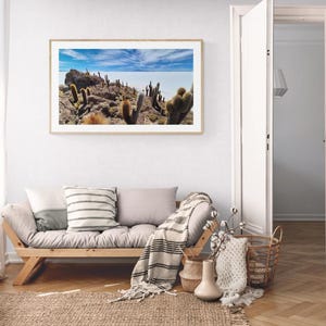 Peut inclure: Une photographie de paysage rectangulaire dans un cadre en bois clair représentant un terrain rocheux avec de grands cactus sous un ciel bleu vif parsemé de nuages fins. L'image montre un paysage désertique avec des rochers bruns.