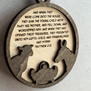 May include: A circular wooden ornament featuring a dark brown laser-cut frame and a light wood center. The center displays the text: AND WHEN THEY WERE COME INTO THE HOUSE, THEY SAW THE YOUNG CHILD WITH MARY HIS MOTHER, AND FELL DOWN, AND WORSHIPPED HIM: AND WHEN THEY HAD OPENED THEIR TREASURES, THEY PRESENTED UNTO HIM GIFTS; GOLD, AND FRANKINCENSE AND MYRRH MATTHEW 2:11. Below the text are dark brown silhouettes of a sheep, a donkey, and a manger.