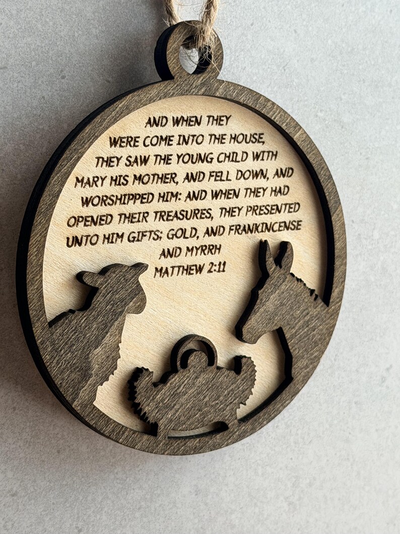 May include: A circular wooden ornament featuring a dark brown laser-cut frame and a light wood center. The center displays the text: AND WHEN THEY WERE COME INTO THE HOUSE, THEY SAW THE YOUNG CHILD WITH MARY HIS MOTHER, AND FELL DOWN, AND WORSHIPPED HIM: AND WHEN THEY HAD OPENED THEIR TREASURES, THEY PRESENTED UNTO HIM GIFTS; GOLD, AND FRANKINCENSE AND MYRRH MATTHEW 2:11. Below the text are dark brown silhouettes of a sheep, a donkey, and a manger.