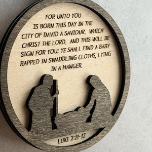 May include: A circular wooden ornament featuring a laser-cut nativity scene silhouette at the bottom. The text reads: FOR UNTO YOU IS BORN THIS DAY IN THE CITY OF DAVID A SAVIOUR, WHICH CHRIST THE LORD. AND THIS WILL BE SIGN FOR YOU; YE SHALL FIND A BABY RAPPED IN SWADDLING CLOTHS, LYING IN A MANGER. LUKE 2:11-12.