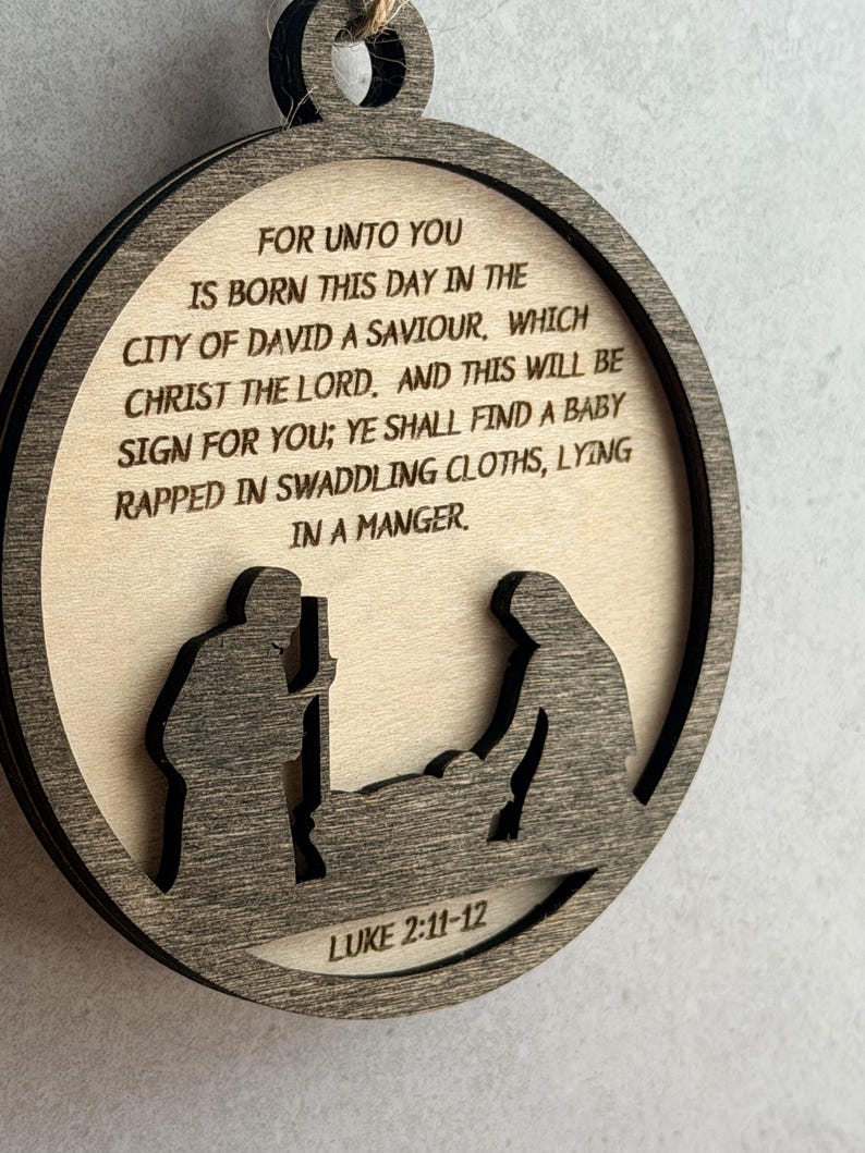 May include: A circular wooden ornament featuring a laser-cut nativity scene silhouette at the bottom. The text reads: FOR UNTO YOU IS BORN THIS DAY IN THE CITY OF DAVID A SAVIOUR, WHICH CHRIST THE LORD. AND THIS WILL BE SIGN FOR YOU; YE SHALL FIND A BABY RAPPED IN SWADDLING CLOTHS, LYING IN A MANGER. LUKE 2:11-12.