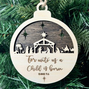 May include: A round wooden Christmas ornament featuring a two-tone design with a dark wood cutout of a nativity scene against a light wood background. The text "For unto us a Child is born" and "ISAIAH 9:6" is engraved on the lower light wood section.
