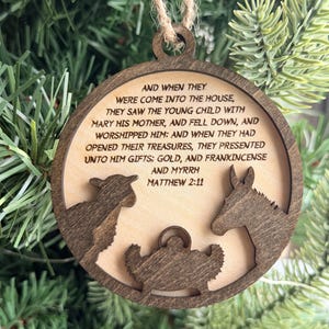 May include: A circular wooden ornament featuring a laser-cut nativity scene with a sheep, a donkey, and a manger. The center is engraved with the text: AND WHEN THEY WERE COME INTO THE HOUSE, THEY SAW THE YOUNG CHILD WITH MARY HIS MOTHER, AND FELL DOWN, AND WORSHIPPED HIM: AND WHEN THEY HAD OPENED THEIR TREASURES, THEY PRESENTED UNTO HIM GIFTS; GOLD, AND FRANKINCENSE AND MYRRH MATTHEW 2:11.