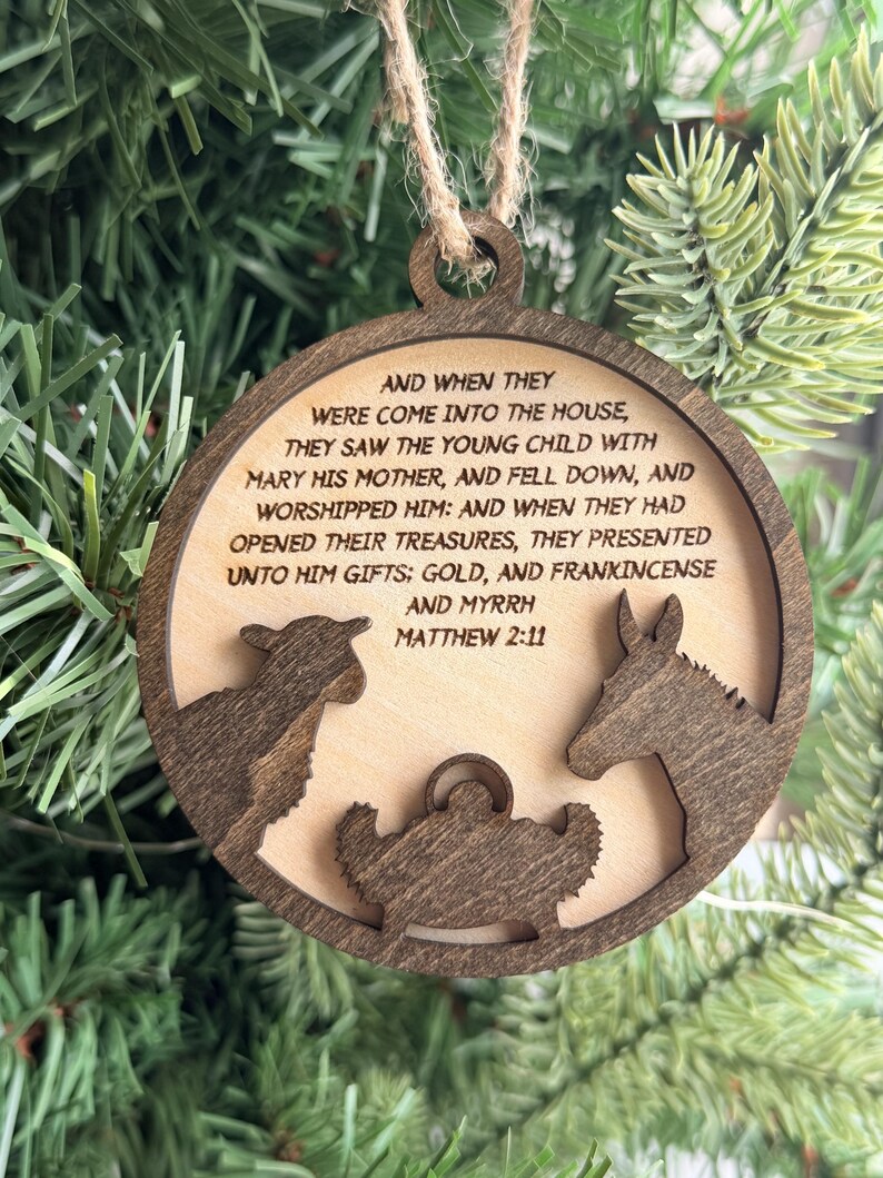 May include: A circular wooden ornament featuring a laser-cut nativity scene with a sheep, a donkey, and a manger. The center is engraved with the text: AND WHEN THEY WERE COME INTO THE HOUSE, THEY SAW THE YOUNG CHILD WITH MARY HIS MOTHER, AND FELL DOWN, AND WORSHIPPED HIM: AND WHEN THEY HAD OPENED THEIR TREASURES, THEY PRESENTED UNTO HIM GIFTS; GOLD, AND FRANKINCENSE AND MYRRH MATTHEW 2:11.