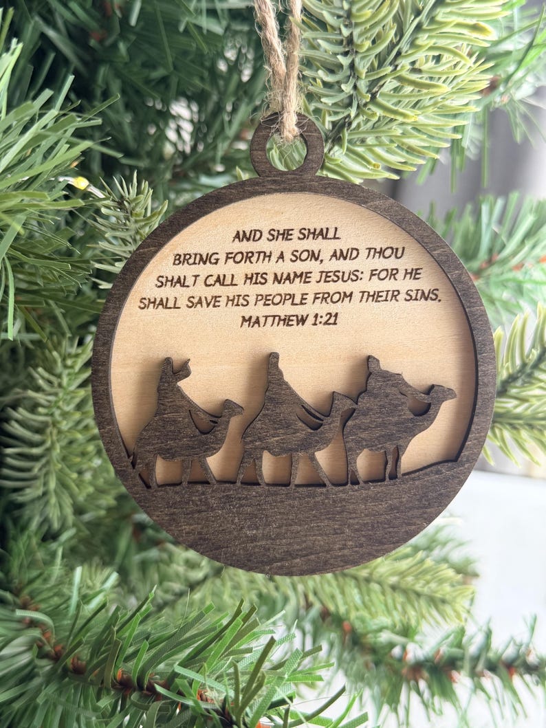 May include: A round wooden ornament featuring a dark brown frame and a light wood center. The text reads: AND SHE SHALL BRING FORTH A SON, AND THOU SHALT CALL HIS NAME JESUS: FOR HE SHALL SAVE HIS PEOPLE FROM THEIR SINS, MATTHEW 1:21. Three camels are below.