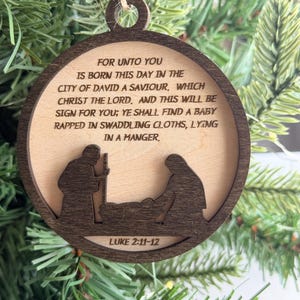 May include: A round wooden Christmas ornament featuring a dark brown frame and a light wood center. The center is engraved with the text: FOR UNTO YOU IS BORN THIS DAY IN THE CITY OF DAVID A SAVIOUR, WHICH CHRIST THE LORD. AND THIS WILL BE SIGN FOR YOU; YE SHALL FIND A BABY RAPPED IN SWADDLING CLOTHS, LYING IN A MANGER. Below the text is a silhouette of a nativity scene and the text LUKE 2:11-12.