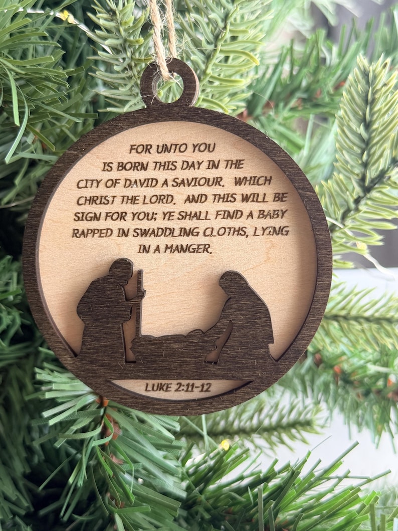 May include: A round wooden Christmas ornament featuring a dark brown frame and a light wood center. The center is engraved with the text: FOR UNTO YOU IS BORN THIS DAY IN THE CITY OF DAVID A SAVIOUR, WHICH CHRIST THE LORD. AND THIS WILL BE SIGN FOR YOU; YE SHALL FIND A BABY RAPPED IN SWADDLING CLOTHS, LYING IN A MANGER. Below the text is a silhouette of a nativity scene and the text LUKE 2:11-12.