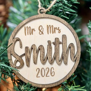 May include: A round wooden Christmas ornament featuring a dark brown stained outer ring and a light wood center. The text "Mr & Mrs" is engraved at the top, the name "Smith" is in raised dark wood lettering, and "2026" is engraved at the bottom.
