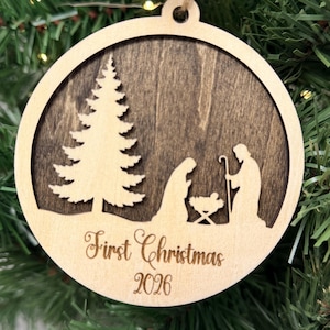 May include: A round wooden Christmas ornament featuring a dark brown stained background with a light-colored wood cutout of a nativity scene and a pine tree. The text First Christmas 2026 is engraved on the bottom of the light wood frame.