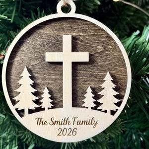 May include: A round wooden ornament featuring a dark brown stained background with a light-colored wooden cross and four pine trees layered on top. The bottom section includes the engraved text The Smith Family and the year 2026.