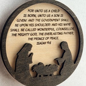 May include: A circular wooden ornament featuring a dark brown frame and a light wood center. The center displays a laser-cut nativity scene silhouette and the text: FOR UNTO US A CHILD IS BORN, UNTO US A SON IS GIVEN: AND THE GOVERNMENT SHALL BE UPON HIS SHOULDER: AND HIS NAME SHALL BE CALLED WONDERFUL, COUNSELLOR, THE MIGHTY GOD, THE EVERLASTING FATHER, THE PRINCE OF PEACE. ISAIAH 9:6.