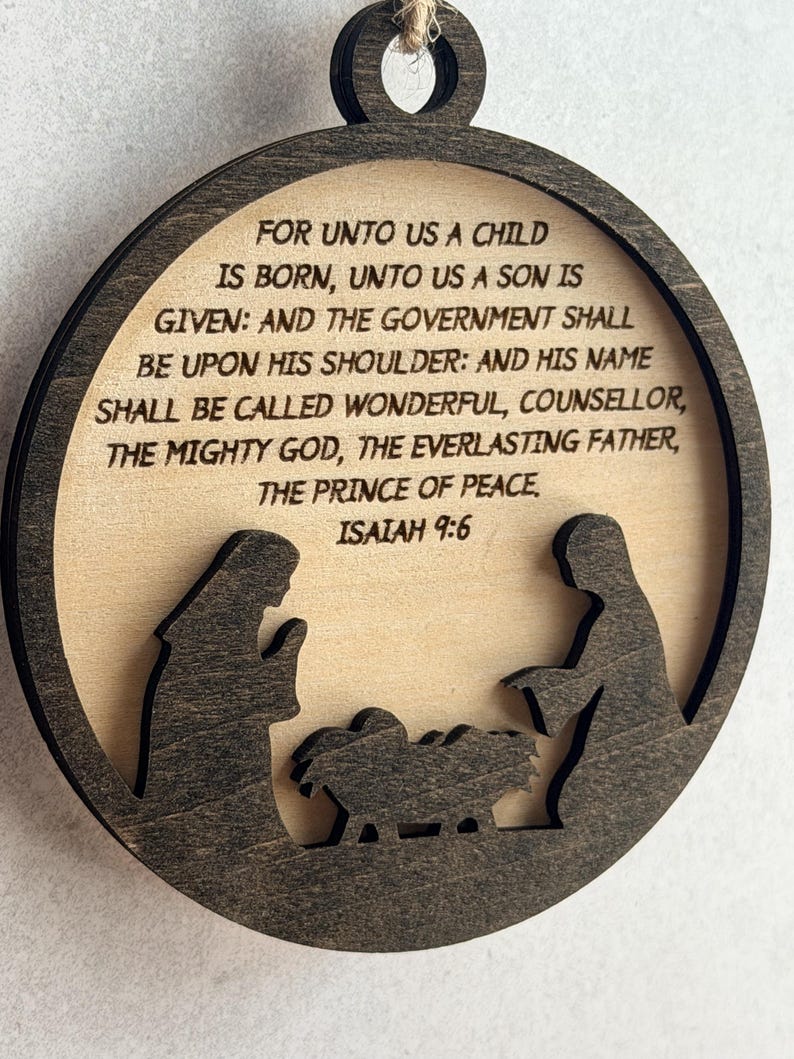 May include: A circular wooden ornament featuring a dark brown frame and a light wood center. The center displays a laser-cut nativity scene silhouette and the text: FOR UNTO US A CHILD IS BORN, UNTO US A SON IS GIVEN: AND THE GOVERNMENT SHALL BE UPON HIS SHOULDER: AND HIS NAME SHALL BE CALLED WONDERFUL, COUNSELLOR, THE MIGHTY GOD, THE EVERLASTING FATHER, THE PRINCE OF PEACE. ISAIAH 9:6.