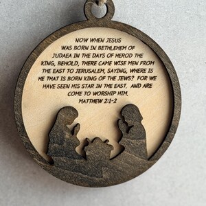 May include: A circular wooden ornament featuring a dark brown frame and a light beige center. The center displays the text: NOW WHEN JESUS WAS BORN IN BETHLEHEM OF JUDAEA IN THE DAYS OF HEROD THE KING, BEHOLD, THERE CAME WISE MEN FROM THE EAST TO JERUSALEM, SAYING, WHERE IS HE THAT IS BORN KING OF THE JEWS? FOR WE HAVE SEEN HIS STAR IN THE EAST. AND ARE COME TO WORSHIP HIM. MATTHEW 2:1-2. Below the text are dark brown silhouettes of two figures kneeling beside a manger.