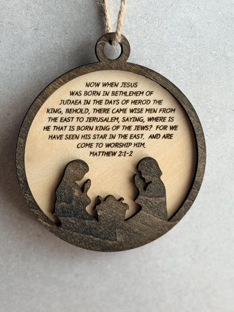 May include: A circular wooden ornament featuring a dark brown frame and a light beige center. The center displays the text: NOW WHEN JESUS WAS BORN IN BETHLEHEM OF JUDAEA IN THE DAYS OF HEROD THE KING, BEHOLD, THERE CAME WISE MEN FROM THE EAST TO JERUSALEM, SAYING, WHERE IS HE THAT IS BORN KING OF THE JEWS? FOR WE HAVE SEEN HIS STAR IN THE EAST. AND ARE COME TO WORSHIP HIM. MATTHEW 2:1-2. Below the text are dark brown silhouettes of two figures kneeling beside a manger.