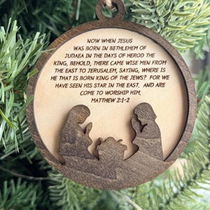 May include: A round wooden ornament featuring a dark brown frame and a light wood center. The center is engraved with the text Matthew 2:1-2 and the story of the nativity. A dark brown silhouette of two figures kneeling by a manger is layered.