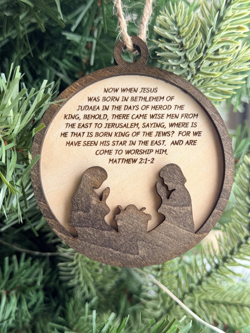 May include: A round wooden ornament featuring a dark brown frame and a light wood center. The center is engraved with the text Matthew 2:1-2 and the story of the nativity. A dark brown silhouette of two figures kneeling by a manger is layered.