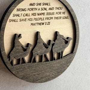 May include: A circular wooden ornament featuring a dark brown frame and a light beige center. The center displays the text: AND SHE SHALL BRING FORTH A SON, AND THOU SHALT CALL HIS NAME JESUS: FOR HE SHALL SAVE HIS PEOPLE FROM THEIR SINS, MATTHEW 1:21.