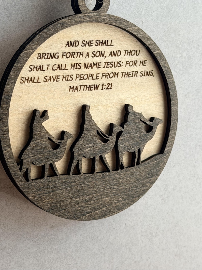 May include: A circular wooden ornament featuring a dark brown frame and a light beige center. The center displays the text: AND SHE SHALL BRING FORTH A SON, AND THOU SHALT CALL HIS NAME JESUS: FOR HE SHALL SAVE HIS PEOPLE FROM THEIR SINS, MATTHEW 1:21.