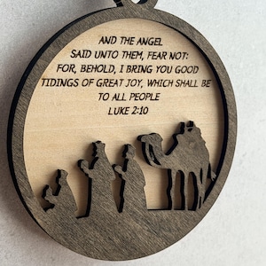 May include: A circular wooden ornament featuring a dark brown frame and a light wood center. The text reads: AND THE ANGEL SAID UNTO THEM, FEAR NOT: FOR, BEHOLD, I BRING YOU GOOD TIDINGS OF GREAT JOY, WHICH SHALL BE TO ALL PEOPLE LUKE 2:10. Below is a silhouette.