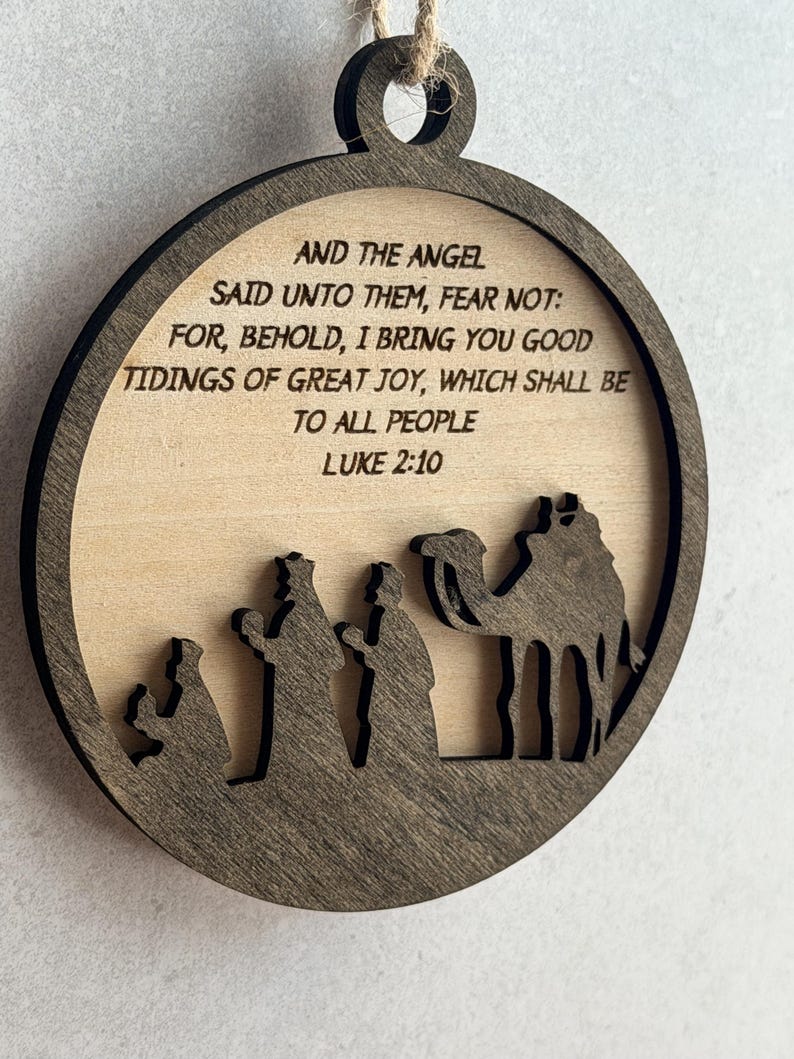 May include: A circular wooden ornament featuring a dark brown frame and a light wood center. The text reads: AND THE ANGEL SAID UNTO THEM, FEAR NOT: FOR, BEHOLD, I BRING YOU GOOD TIDINGS OF GREAT JOY, WHICH SHALL BE TO ALL PEOPLE LUKE 2:10. Below is a silhouette.