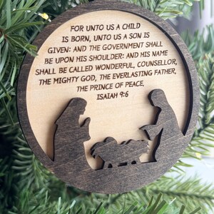May include: A circular wooden ornament featuring a dark brown frame and a light wood center. The center is engraved with the text: FOR UNTO US A CHILD IS BORN, UNTO US A SON IS GIVEN: AND THE GOVERNMENT SHALL BE UPON HIS SHOULDER: AND HIS NAME SHALL BE CALLED WONDERFUL, COUNSELLOR, THE MIGHTY GOD, THE EVERLASTING FATHER, THE PRINCE OF PEACE. ISAIAH 9:6. Below the text is a dark brown silhouette of a nativity scene.
