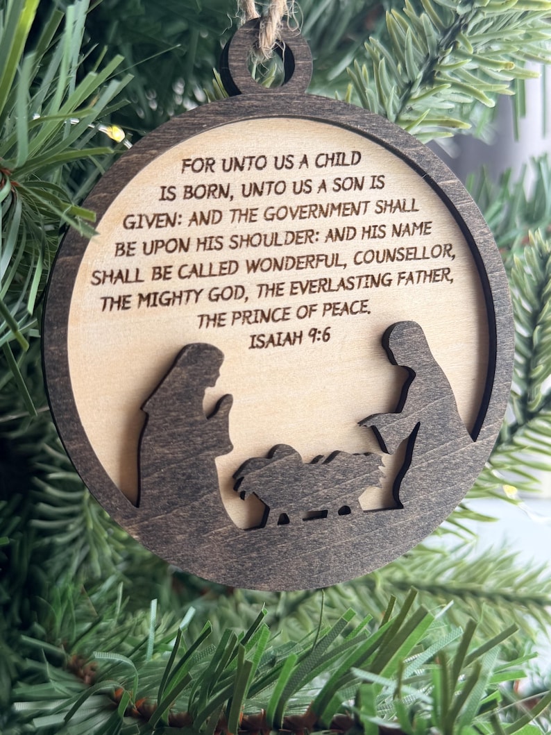 May include: A circular wooden ornament featuring a dark brown frame and a light wood center. The center is engraved with the text: FOR UNTO US A CHILD IS BORN, UNTO US A SON IS GIVEN: AND THE GOVERNMENT SHALL BE UPON HIS SHOULDER: AND HIS NAME SHALL BE CALLED WONDERFUL, COUNSELLOR, THE MIGHTY GOD, THE EVERLASTING FATHER, THE PRINCE OF PEACE. ISAIAH 9:6. Below the text is a dark brown silhouette of a nativity scene.